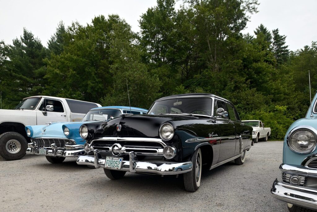 A row of classic cars parked on a gravel lot with lush green trees in the background, featuring a prominent black 1952 Ford Custom in the center.