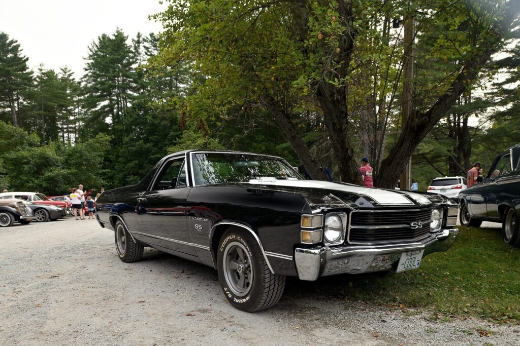 A black and white 1970 Chevrolet El Camino SS parked on a gravel lot with trees and people in the background.