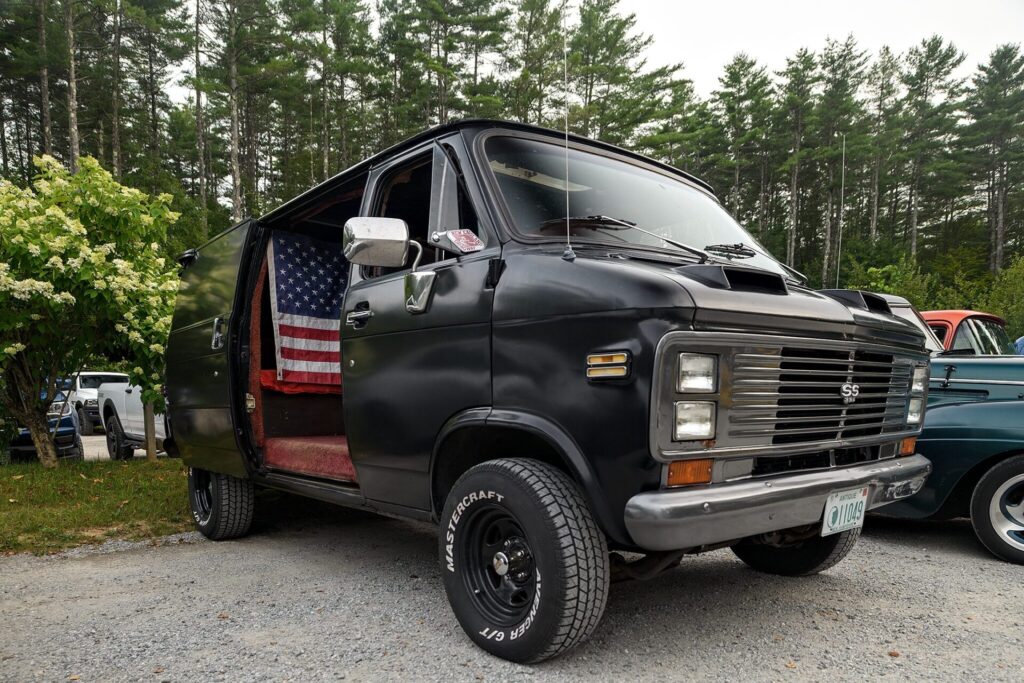 A black 1970s Chevrolet van with an American flag inside the open door, parked on a gravel lot with trees in the background.