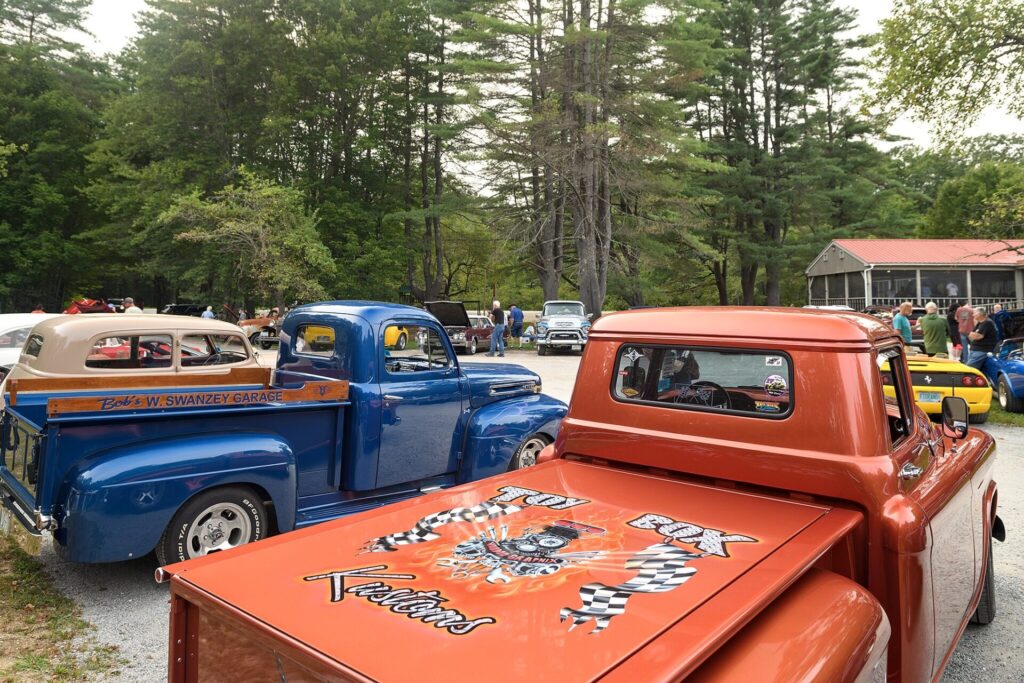 A vibrant orange pickup truck with custom artwork on the tailgate, parked next to a blue truck at a car show with trees and people in the background.