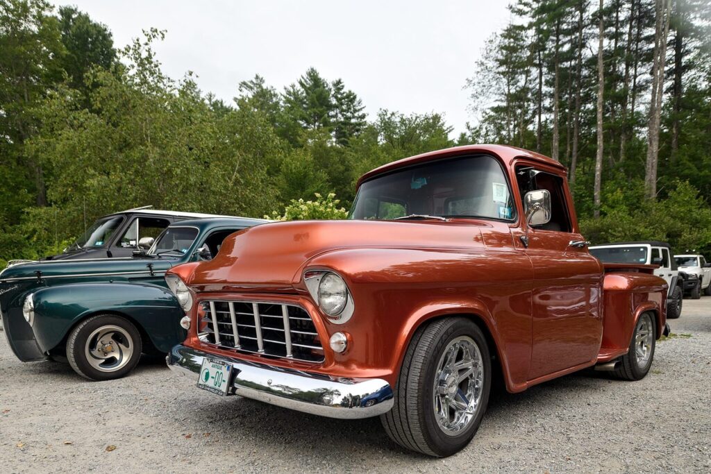 A shiny orange vintage Chevrolet pickup truck parked on a gravel lot with a green classic car beside it and trees in the background.