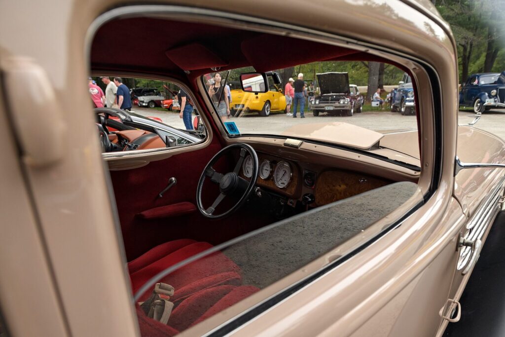Interior view of a beige vintage car with red upholstery, showing the steering wheel and dashboard, with a car show scene visible through the windshield.