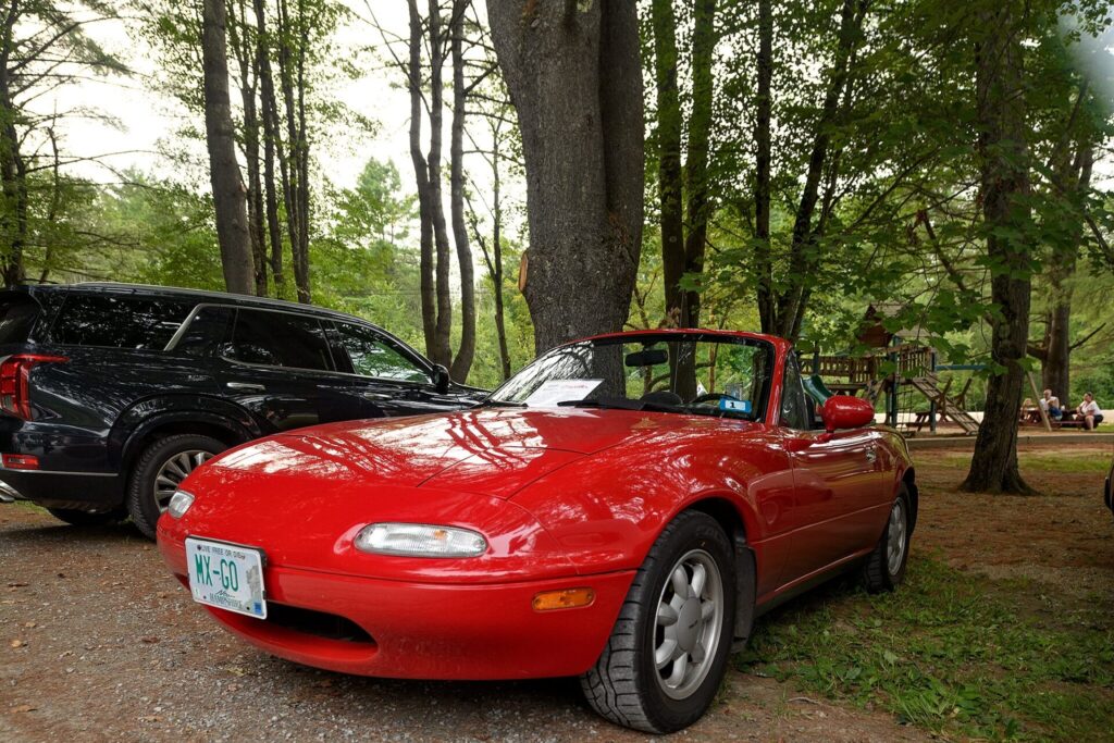 A red Mazda MX-5 Miata convertible parked on a gravel lot with trees and a playground in the background.