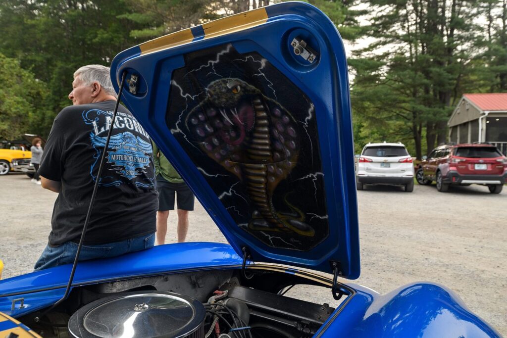 A person sitting on the edge of a blue Shelby Cobra replica with its hood open, revealing a detailed cobra painting inside. The car is parked in a lot with trees and other vehicles in the background. The person is wearing a black shirt with "Laconia Motorcycle Rally" text.