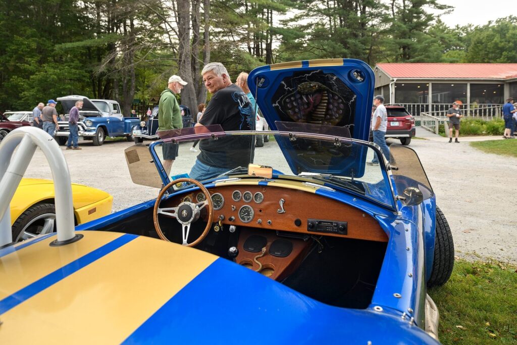 A blue and yellow Shelby Cobra replica with its hood and door open, displayed at an outdoor car show. The car's interior features a wooden dashboard with gauges and a steering wheel, while people gather around other classic vehicles in the background.