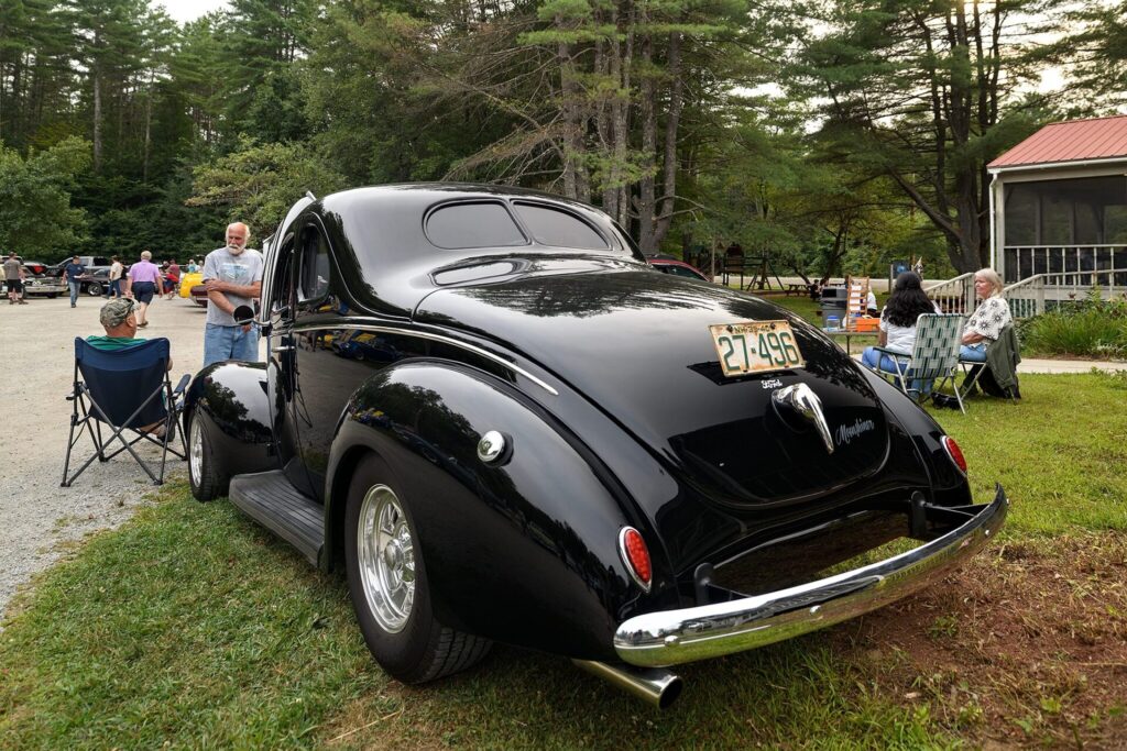 A black vintage car with a license plate reading "27-496" is parked on grass near a gravel path at an outdoor car show. People are seated in chairs and standing around, with trees and a small building in the background.