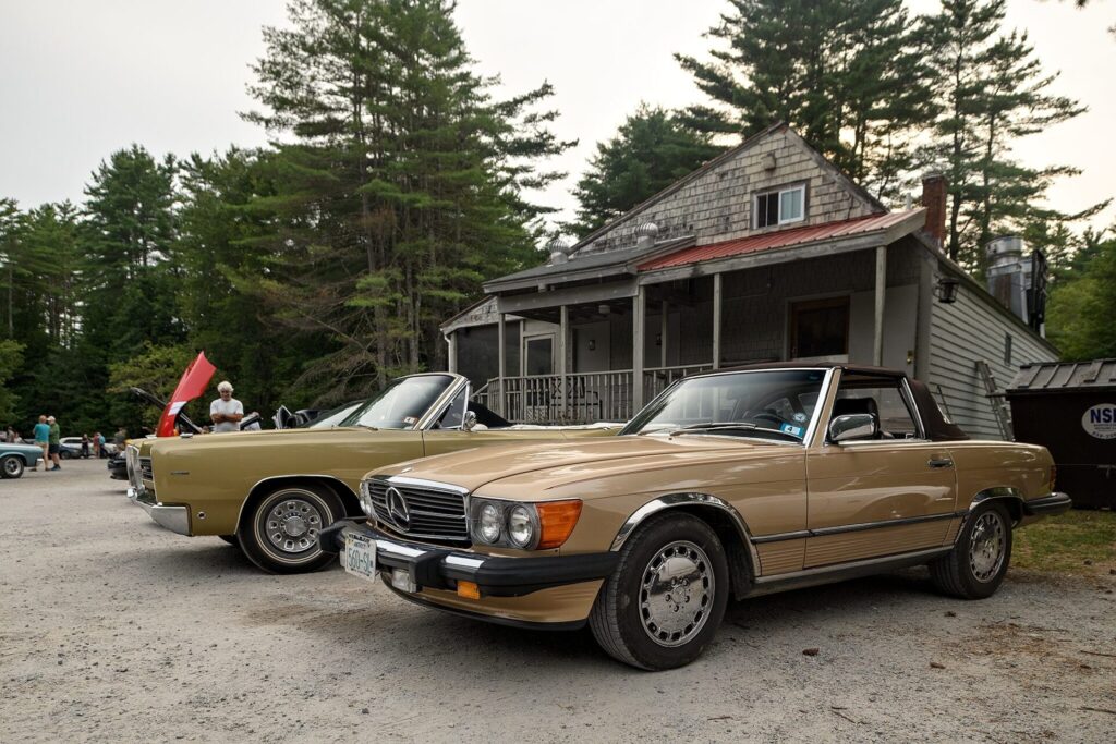 A photo featuring two vintage cars, a beige Mercedes-Benz SL convertible and a green classic car, parked in front of a rustic wooden house with a red roof, surrounded by tall pine trees.