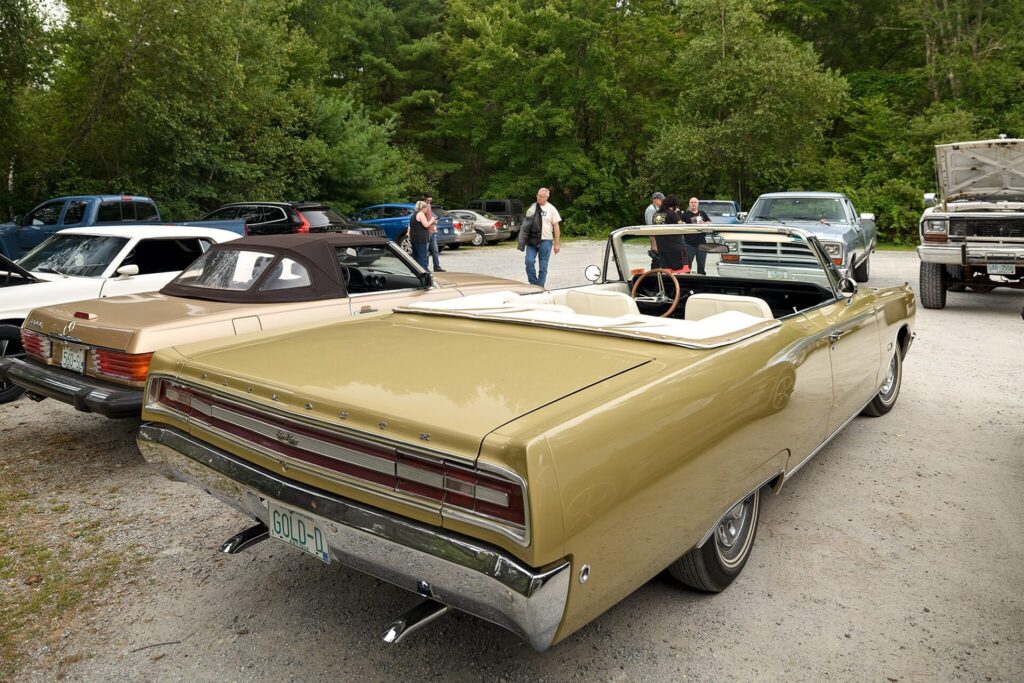 A vintage gold convertible car with its top down, parked on a gravel lot surrounded by trees. Other classic cars and people are visible in the background.