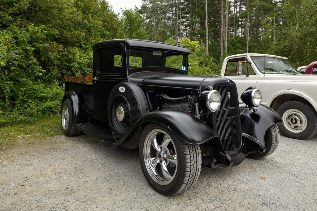 A black vintage Ford truck with a polished chrome grille and wheels, parked on a gravel surface with green trees and foliage in the background. Another older truck is partially visible to the right.