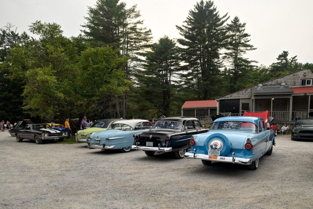 A photo of a car show featuring several classic cars parked on a gravel lot, with a forested background and a rustic building with a red roof.